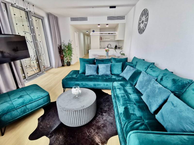 Living room of a 3-room apartment with a turquoise sofa, wooden decor flooring, and a kitchen.
