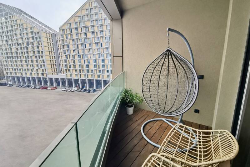 Balcony of a 3-room apartment with a wooden decor floor, a hanging chair, and a view of apartment buildings.