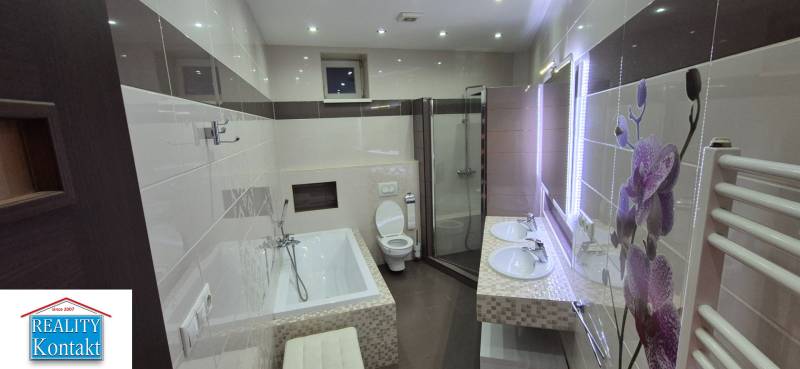 A bathroom in a family house with sinks, a bathtub, a shower, and a lighted mirror.