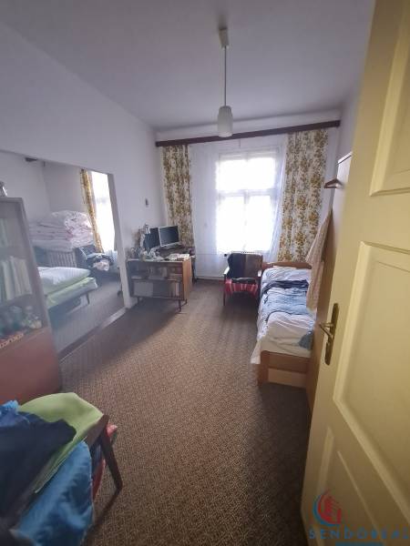 A room in a family house with a carpet, a bed, a computer, and a window with floral curtains.