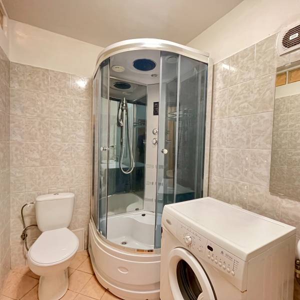 Bathroom in a studio apartment with a corner shower, washing machine, and toilet.