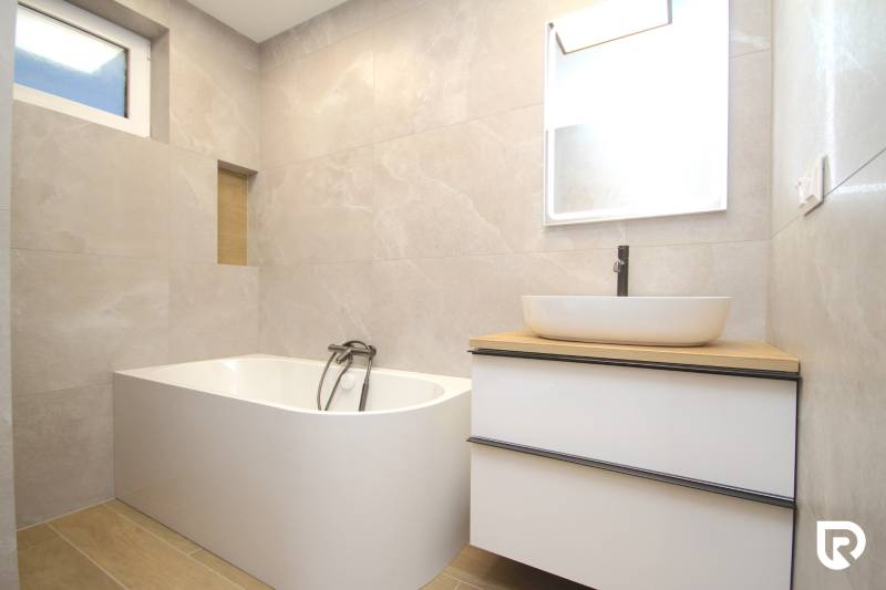 A bathroom in a family house with a bathtub, a sink, a mirror, and a floor with a wooden decor.
