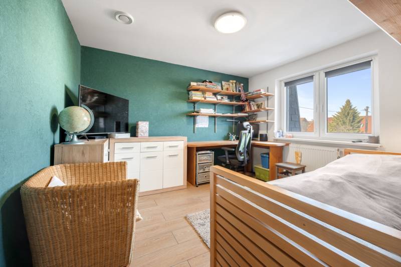 A room in a family house with a desk, a green wall, a television, and a wooden-patterned floor.