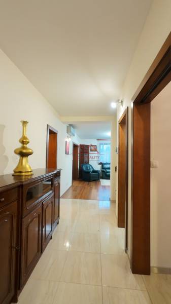 A hallway in a family house with tiles and wooden furniture leads to a living room with a wood-patterned floor.