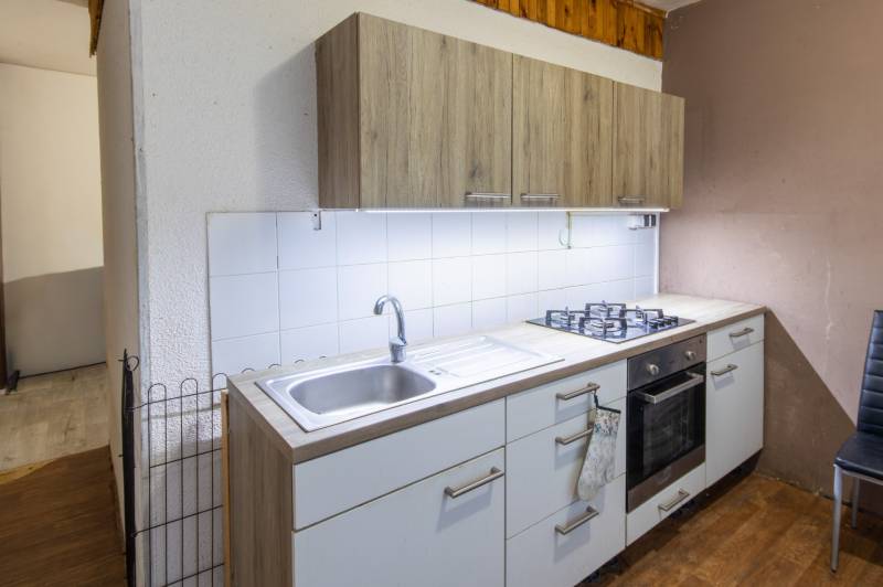 A kitchen with wood-patterned flooring, white tiles, and a gas stove in a 2-room apartment.