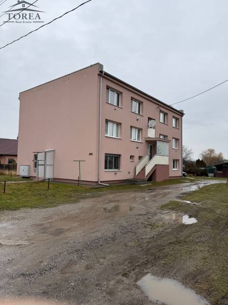 Apartment building in Ludanice with four windows and an entrance staircase on a wet road.