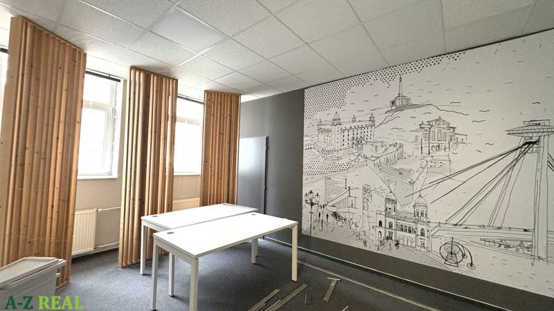 The office has white desks, a drawing on the wall, and vertical window blinds.