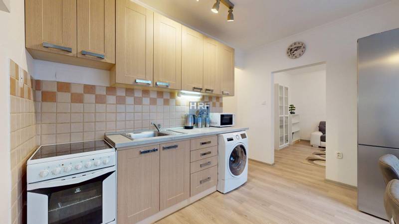 A kitchen in a 2-room apartment with beige cabinets, appliances, and a wooden decor floor.