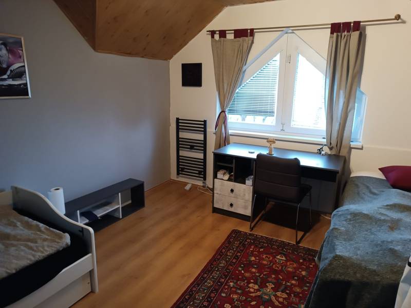 A room in a family house with a wooden decor floor, a desk, a bed, and a carpet.