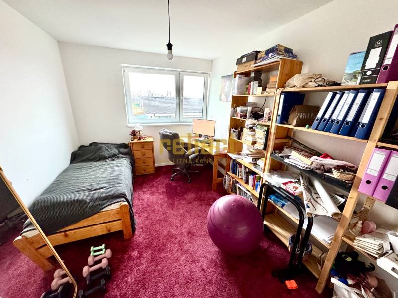 A room in a family house with a table, bed, shelves, and a red carpet.