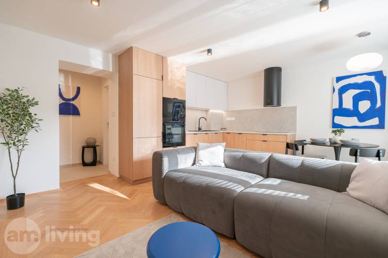 Living area of a 3-room apartment with a kitchen, gray sofa, and wood-patterned flooring.
