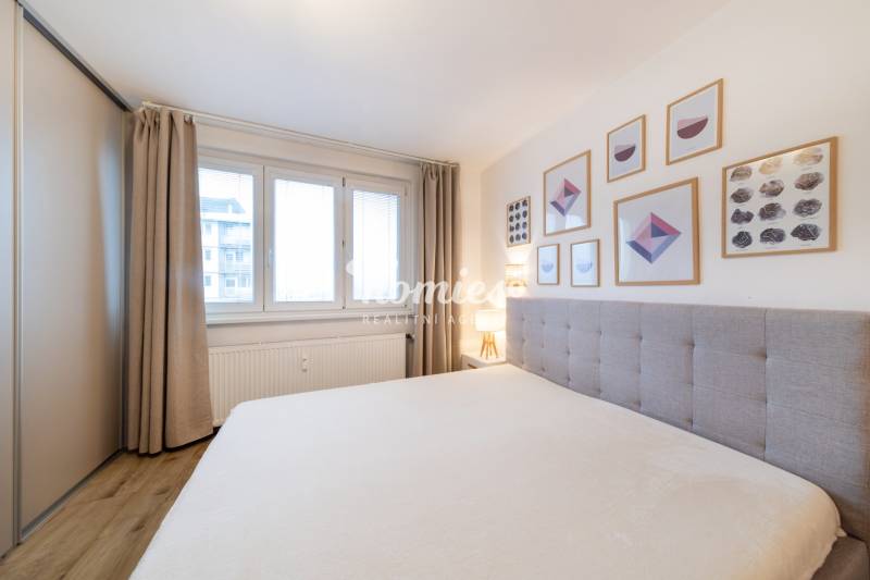 A bright bedroom with wood-patterned flooring, a bed, and wall pictures in a two-room apartment.