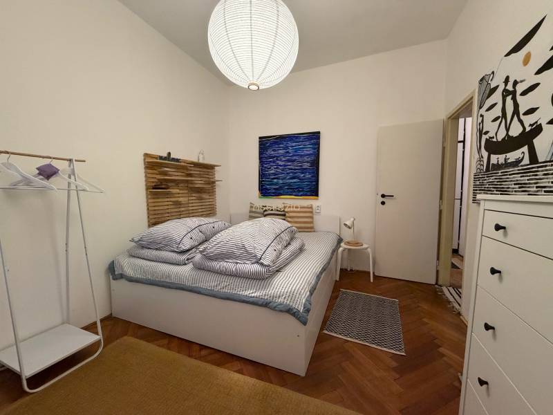 Bedroom in a two-room apartment with wood-patterned flooring, a bed, and a white cabinet.