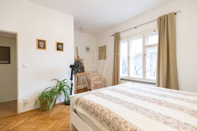 A bright bedroom with wood-patterned flooring, plants, and large windows in a two-room apartment.