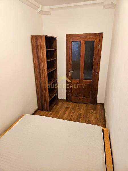 A room in a 3-room apartment with a wooden decor floor, a bed, and a bookshelf.