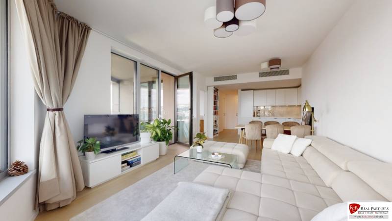 Living room of a three-room apartment with a sofa, TV, light curtains, and wood-patterned flooring.