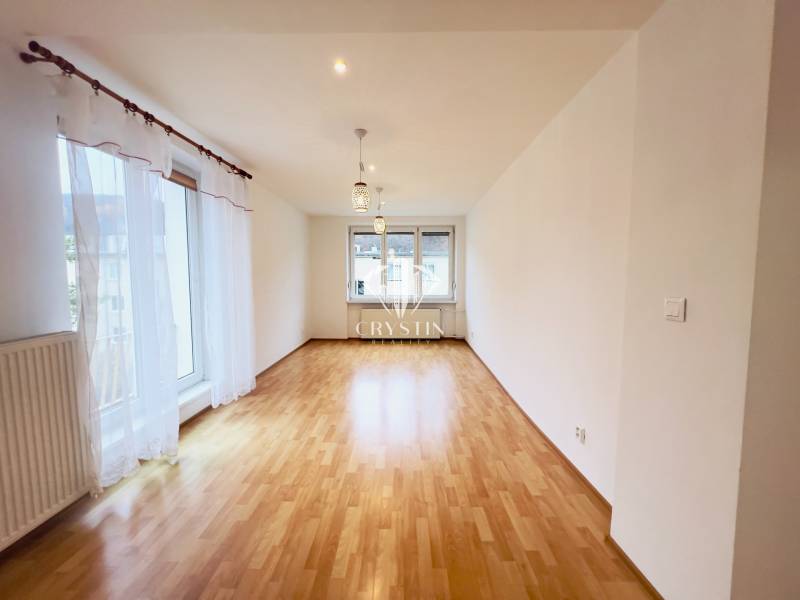 A bright living room in a two-room apartment with wood-patterned flooring and large windows.