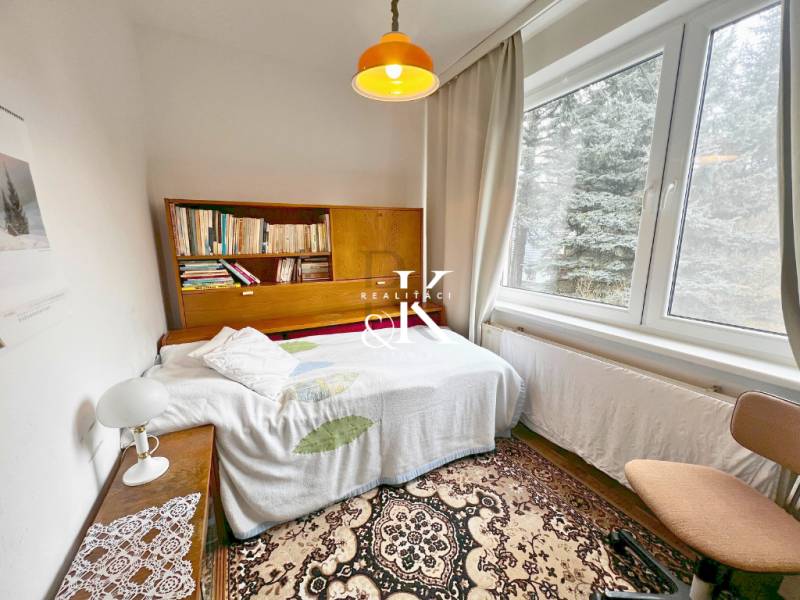 A room in a cottage with a bookshelf, a bed, and a large window.