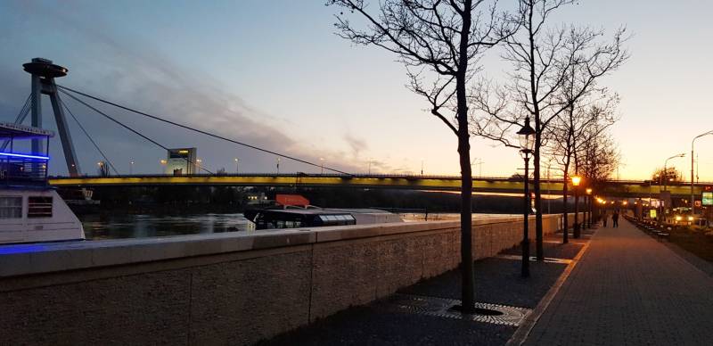 An evening view of the UFO Bridge from Rázus Embankment in Bratislava, with an illuminated walkway.