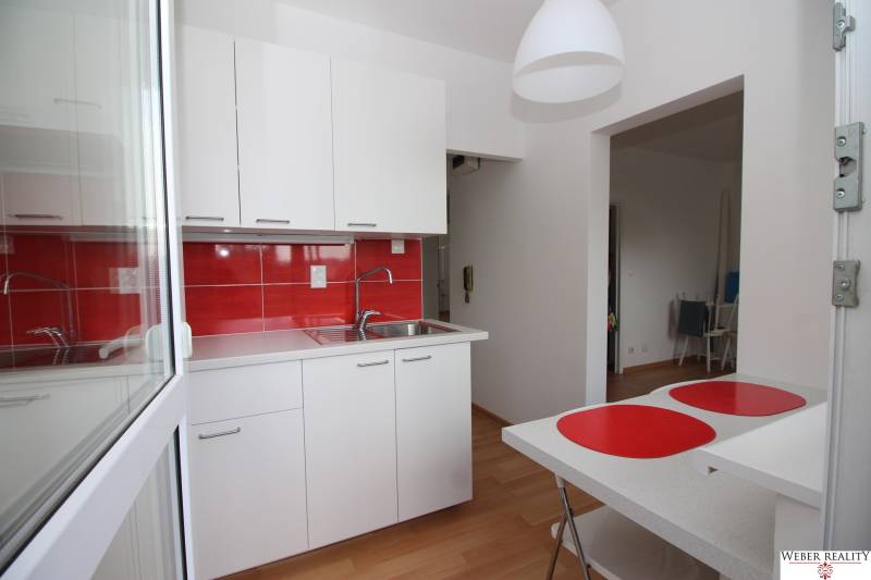 A kitchen with red tiles and white cabinets in a studio apartment, with a wood-patterned floor.