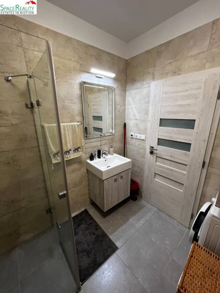 A bathroom in a 2-room apartment with a sink, shower, and washing machine on tiled flooring.