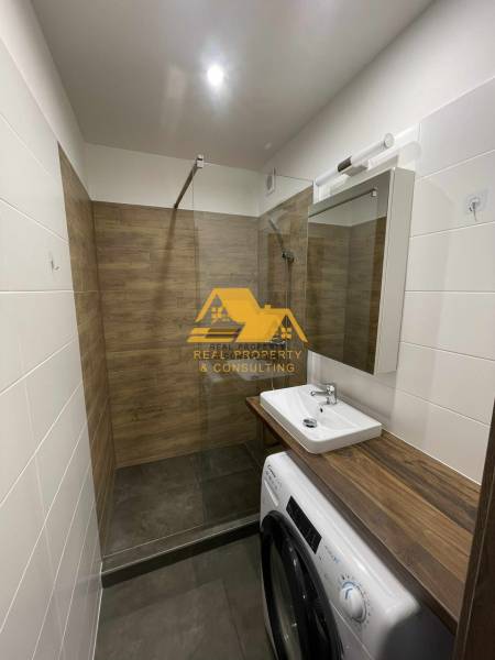 Bathroom in a studio apartment with a sink, shower, and washing machine, wooden decor.