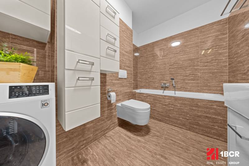 A bathroom in a 2-room apartment with brown tiles, a bathtub, a washing machine, and storage cabinets.