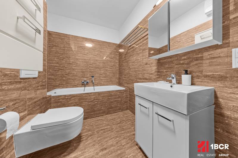 Bathroom with a bathtub and a wall-hung toilet in a 2-room apartment with light tiles.