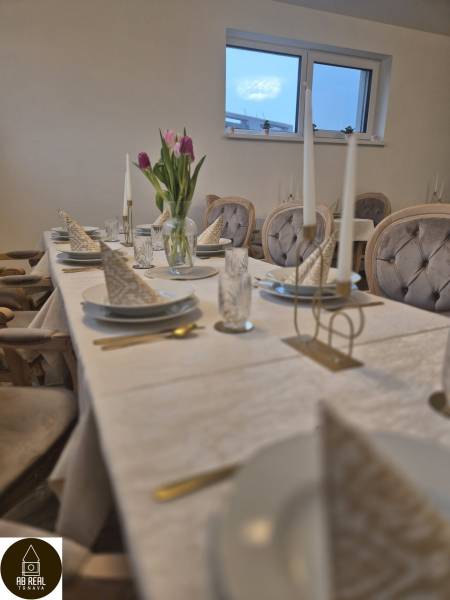 A table with a white tablecloth, plates, glasses, flowers, and chair covers in a dining area.
