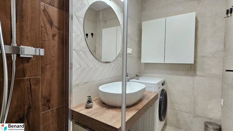 Bathroom in a studio apartment with a sink, shower, and washing machine, floor with wood decor.
