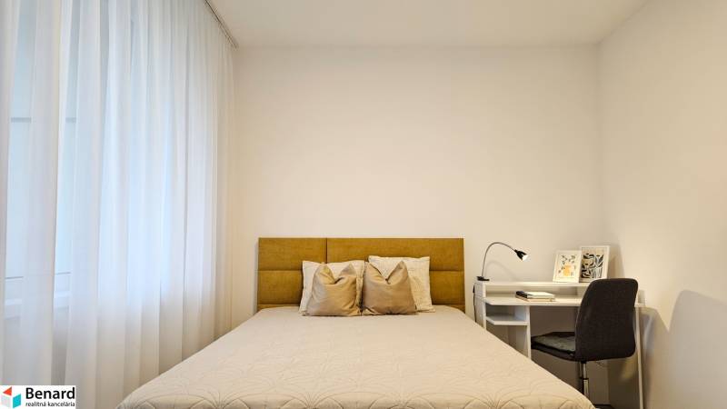 A bedroom in a studio apartment with a light-colored bed, curtains, and a white table with a lamp.