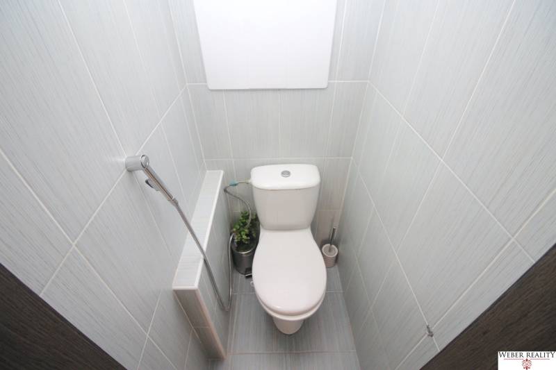 A toilet in a 3-room apartment with beige tiles and a detail of a shower head.