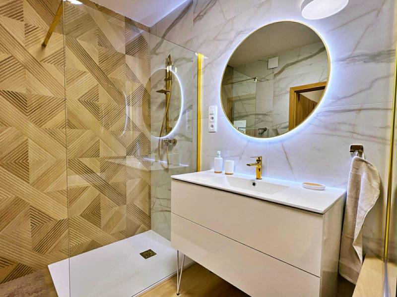 A bathroom in a 3-room apartment with a large illuminated mirror and gold accessories by the shower corner.