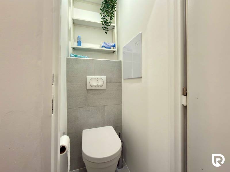 Bathroom in a 2-room apartment with a toilet, white walls, and shelves.