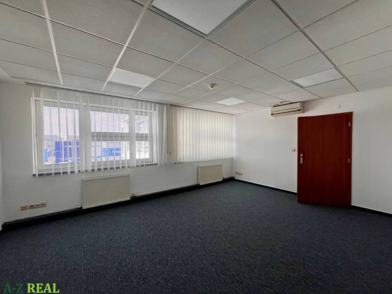 An office with carpeted flooring, white walls, air conditioning, and vertical blinds on the windows.