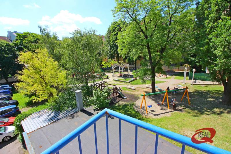 A children's playground surrounded by trees, a parking lot with cars, blue railing.