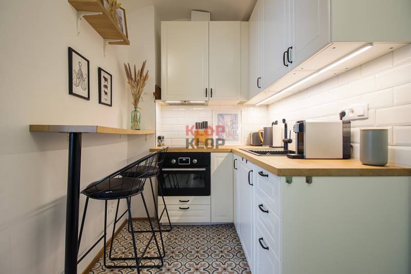 A kitchenette of a one-room apartment with white cabinets, patterned floor, and wooden shelves.