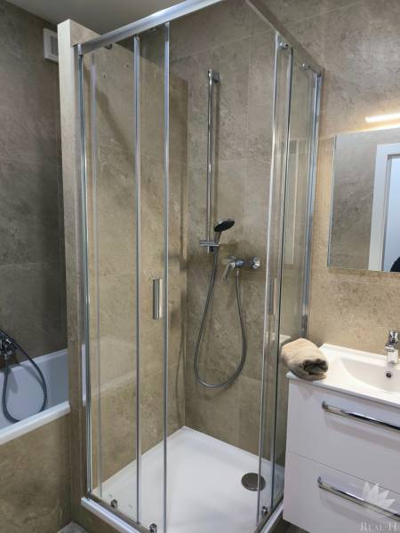 A shower corner with marble tiles in a 4-room apartment, next to a sink with a white cabinet.