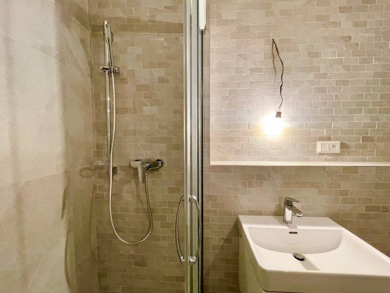 Bathroom in a 2-room apartment with a sink, shower, and light on beige tiles.