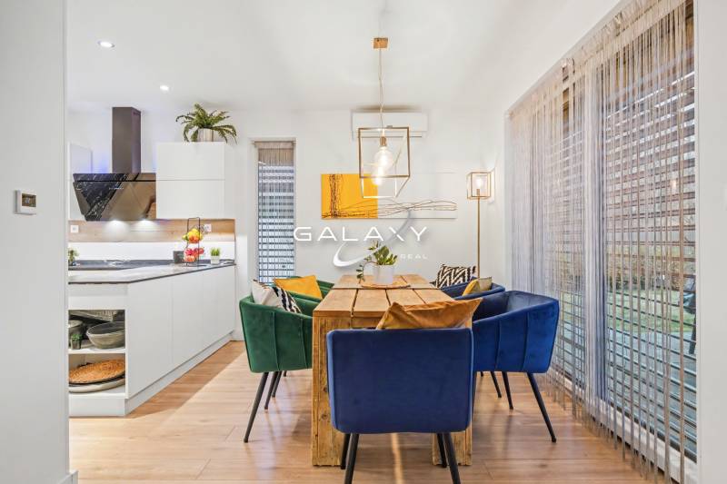 The dining room of a family house with a wooden table, colorful chairs, and a kitchen in the background.