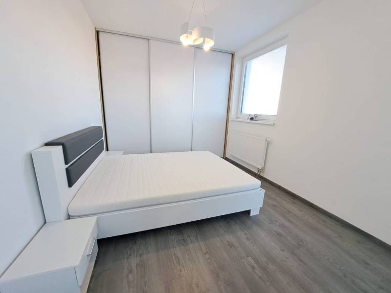 A bright bedroom in a two-room apartment with wood-patterned flooring, white furniture, and a built-in wardrobe.