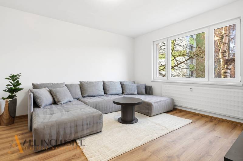 A bright living room with wood-patterned flooring, a gray sofa, and a window in a three-room apartment.