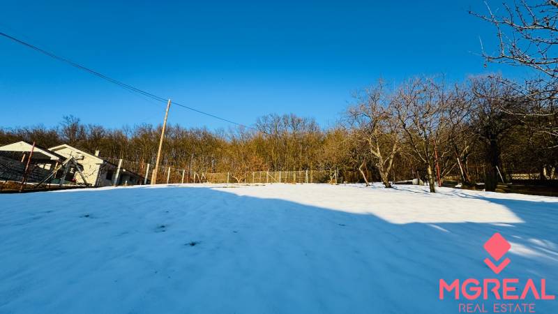 A snowy plot in Horna Ves surrounded by trees and a fence near a family house.