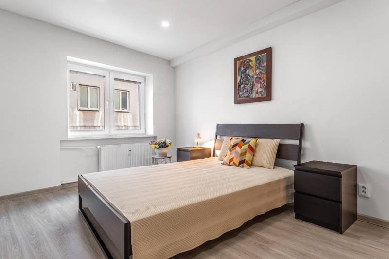 Bedroom in a three-room apartment with a bed, colorful pillows, and a wooden-patterned floor.