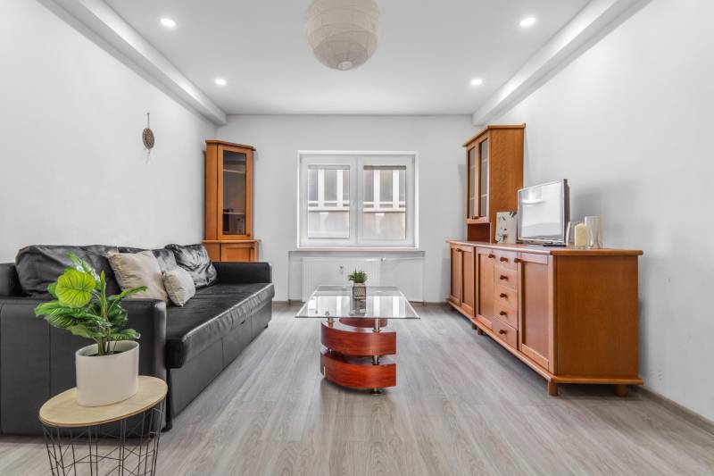 Living room with a black sofa, wooden furniture, and a floor with a wooden decor in a 3-room apartment.