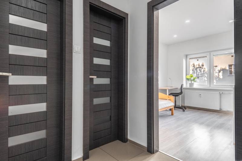 A hallway with dark doors and a view into a room with wood-patterned flooring.