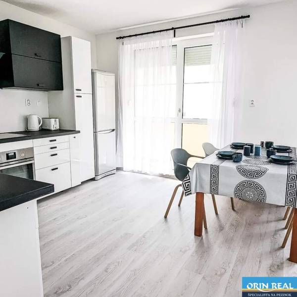 A bright kitchen with a dining table, white cabinetry, and a wood-patterned floor in a two-room apartment.
