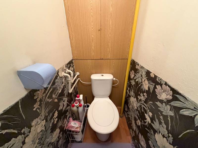 A toilet in a 3-room apartment with floral wallpaper and storage space above the toilet.