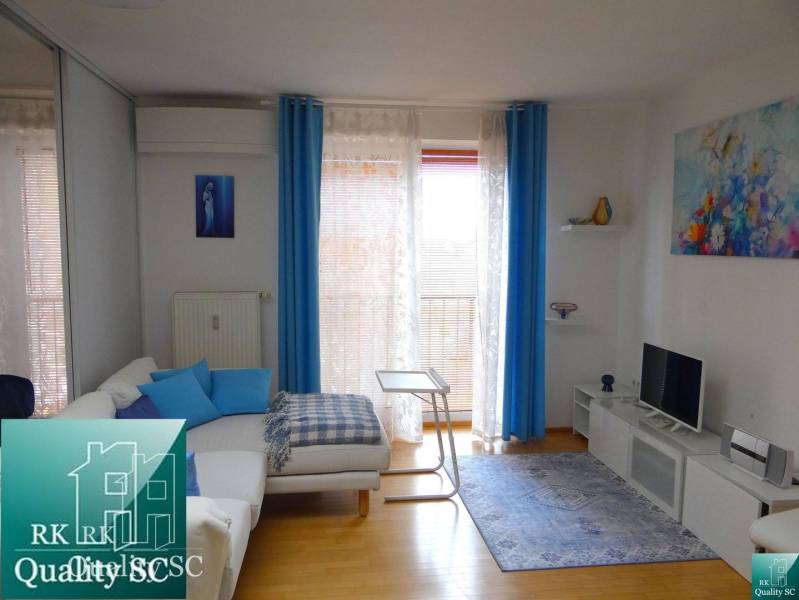 Living room in a studio apartment with a white sofa, blue curtains, and a wooden decor floor.