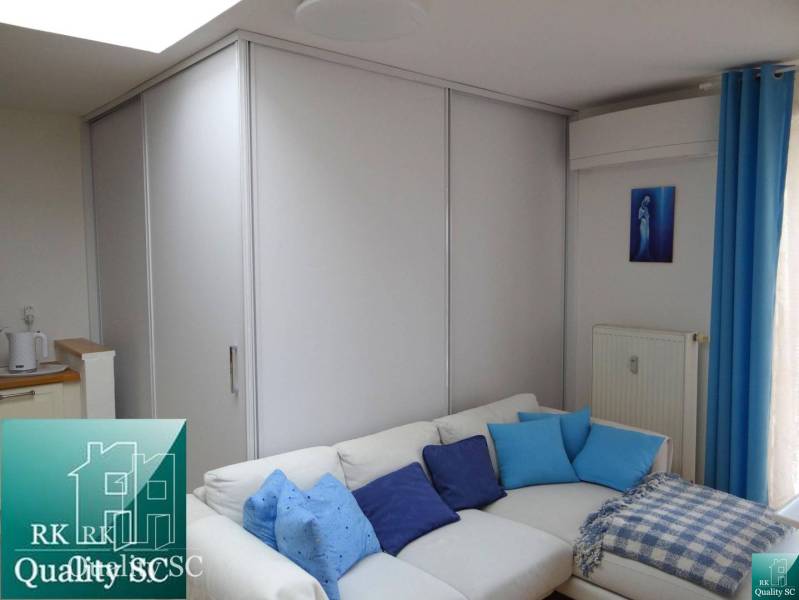A sofa with blue cushions in a studio apartment, next to a wardrobe and curtains.